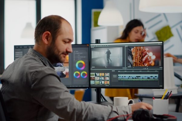 Video maker editing movie using post production software working in creative start up agency office with two monitors. Videographer processing audio film montage on professional computer, taking notes